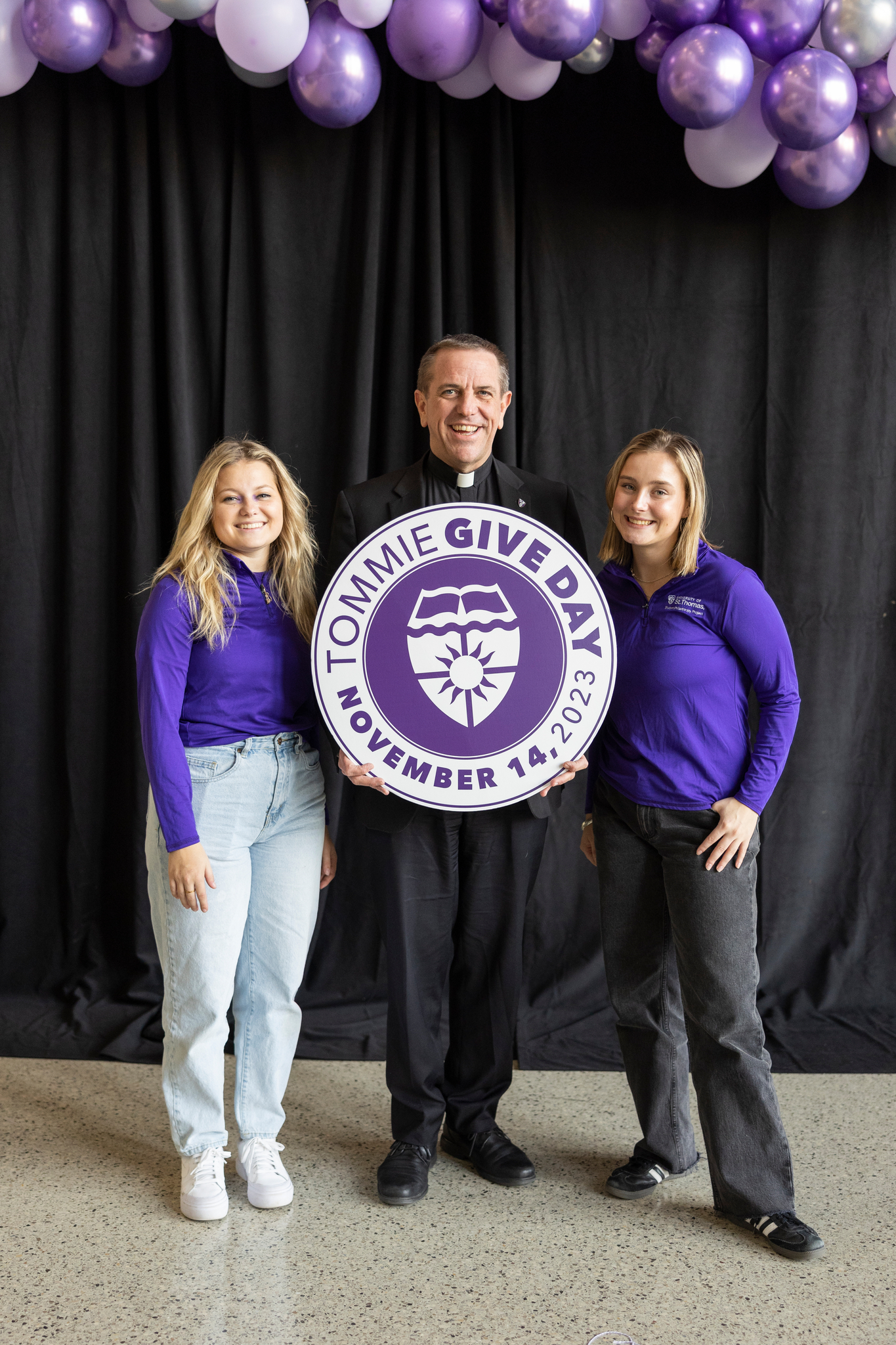 Tommie students and Father Chris Collins holding a Tommie Give Day sign and smiling
