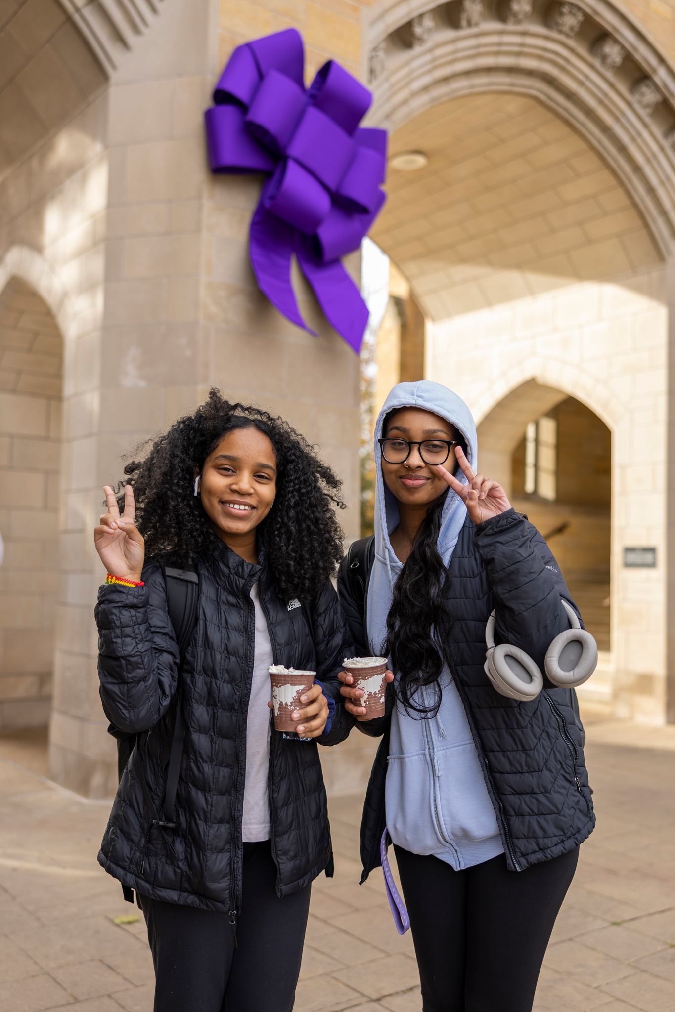 Two students holding hot chocolate in front of a big purple bow hung on The Arches