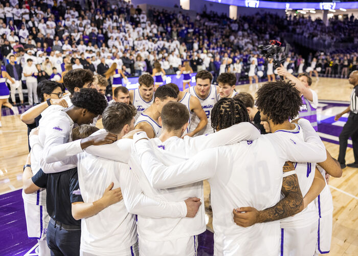 Tommie men's basketball players gather in a huddle on the court before a game as fans fill the arena