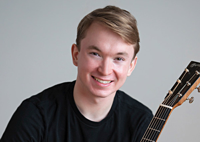 Portrait photo of student Benjamin Barrett. He's holding his guitar against a grey background.