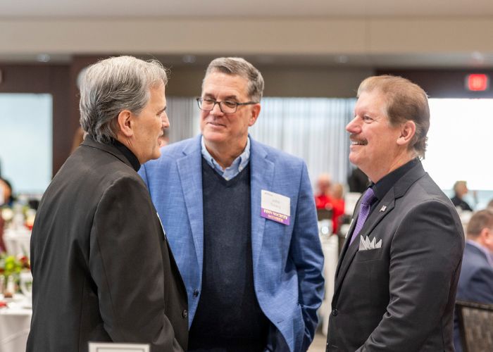 Three Alumni & Friends Christmas Luncheon attendees smile and talk at the 2025 event in Woulfe Alumni Hall. The man in the center wears a nametag with a Finn Legacy Society ribbon attached.