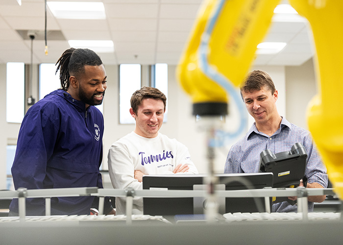 Two students and professor work in an engineering lab; a robotic arm is in the foreground