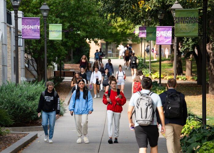 Students walk along a tree-lined campus pathway between classes beneath banners highlighting St. Thomas values