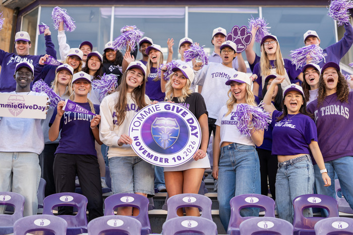 A group of students cheering in the stadium holding a Tommie Give Day sign and wearing purple and white