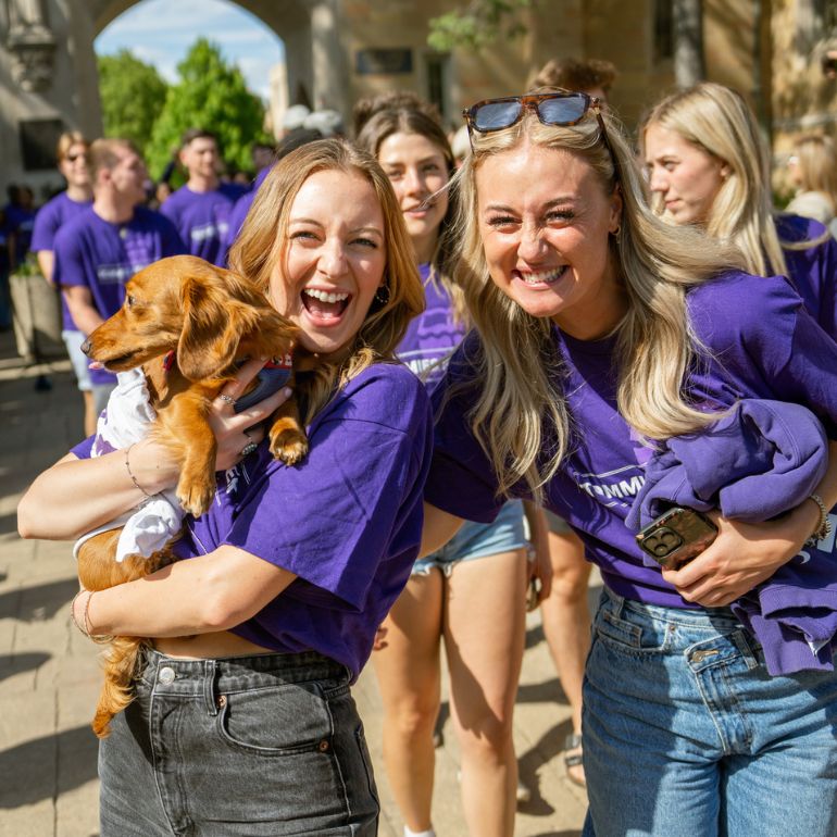 Two smiling students in matching purple tees hold a puppy outside the arches