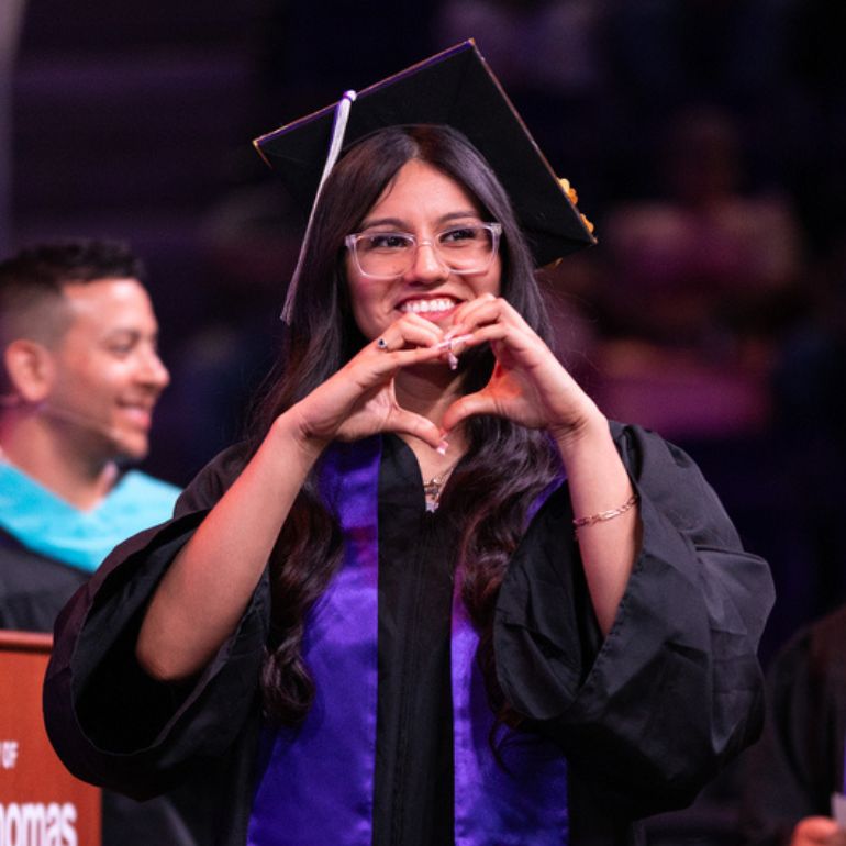 A student making a heart shape with her hands walks across the stage at commencement