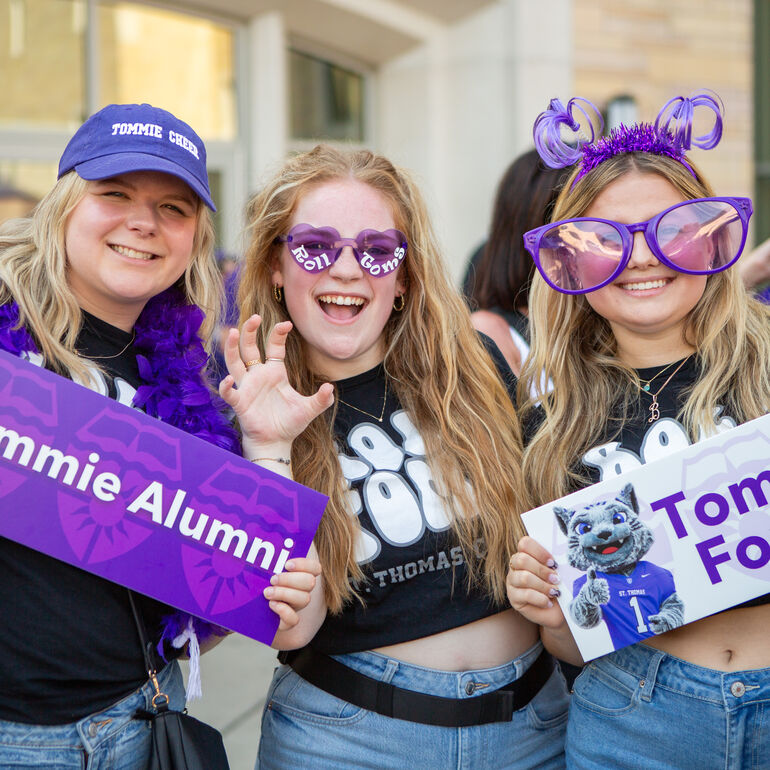 Three people smile while holding "Tommie Alumni" and "Tommies Forever" signs at a St. Thomas celebration.