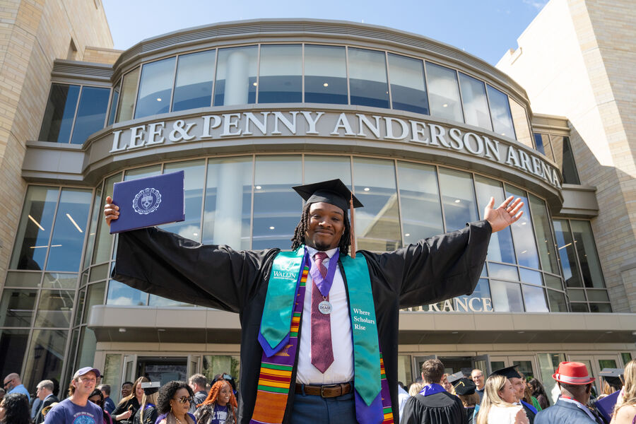 A graduate wearing a graduation gown and mortar board, hold a his arms outstretched with a diploma in one hand in front of a group of graduates and their families walking out of the Lee and Penny Anderson Arena.