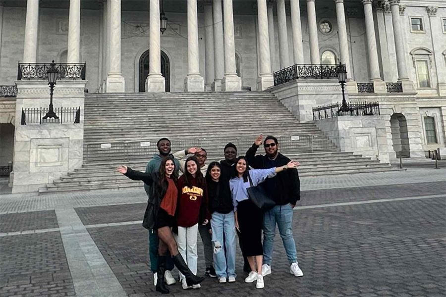 A group of eight people stands in front of the Capitol in Baltimore. They are dressed casually, with some wearing university apparel, and are smiling and waving at the camera.