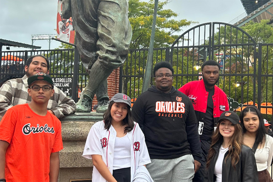 Seven RM&I students pose outside of the Orioles baseball stadium. They’re wearing black and orange Orioles apparel and smiling.
