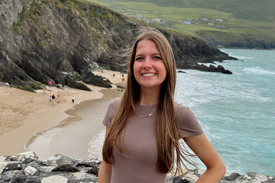 Lauren smiles at the camera, posing in front of a picturesque cliff and a beach below