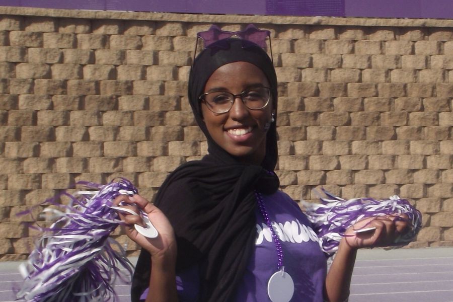 Sukayna smiles into the camera and holds purple pom poms while standing on a St. Thomas athletic field
