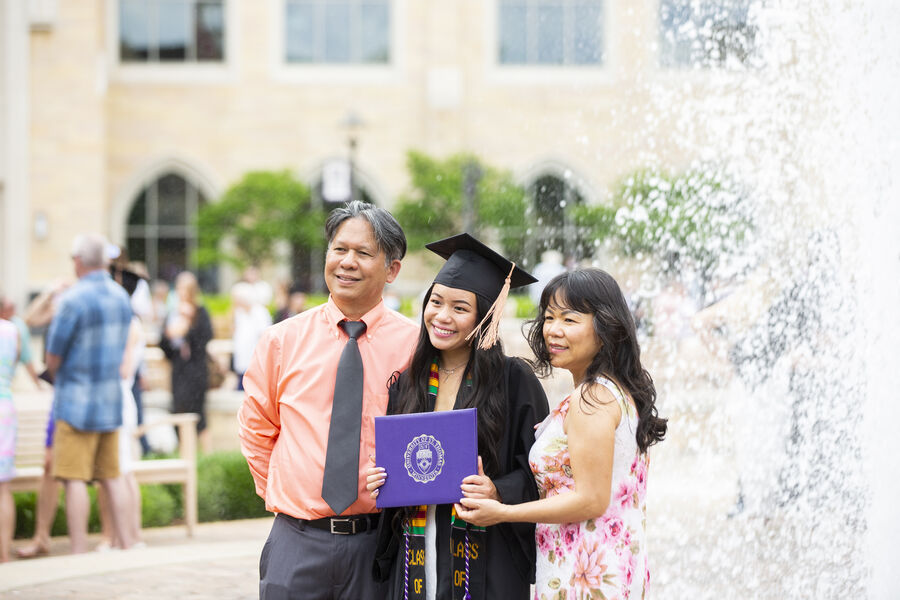 Graduate in cap and gown and holding diploma poses with parents in front of fountain on St. Paul campus. 