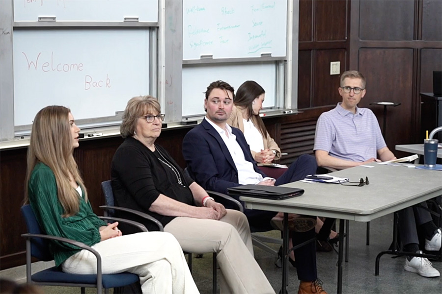 A panel of five people sits at the front of a classroom, engaged in discussion. The room has whiteboards with handwritten notes and 'Welcome Back' written in red. A man in a suit and a woman in green are among the panelists, while another man in glasses and a polo sits at a table.
