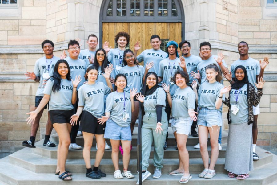 Real Program participants wear matching gray shirts and pose for a group picture on steps outside a campus building