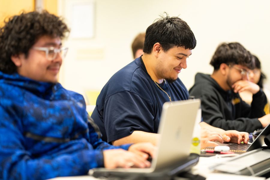 Students participate in a Justice and Peace Studies class at the Dougherty Family College on April 2, 2025, in Minneapolis. Three male students are smiling and studying on laptops.