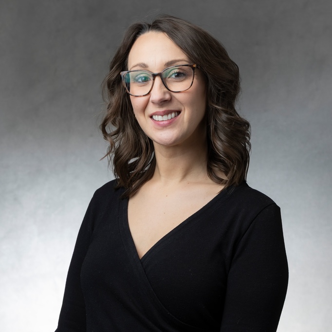Emily Halstead smiles into the camera for a studio portrait taken against a gray background.