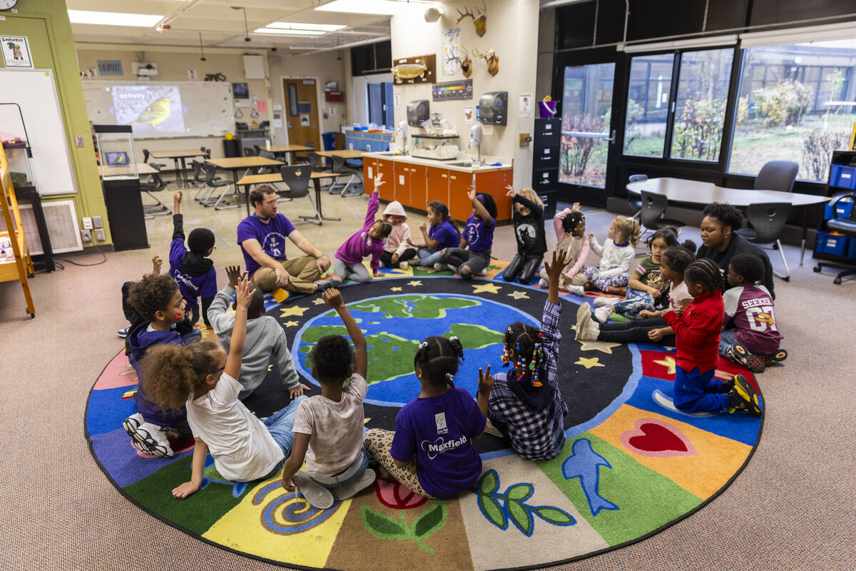 A group of elementary school students sit in a circle on a carpet with their teachers, several students have their hands raised