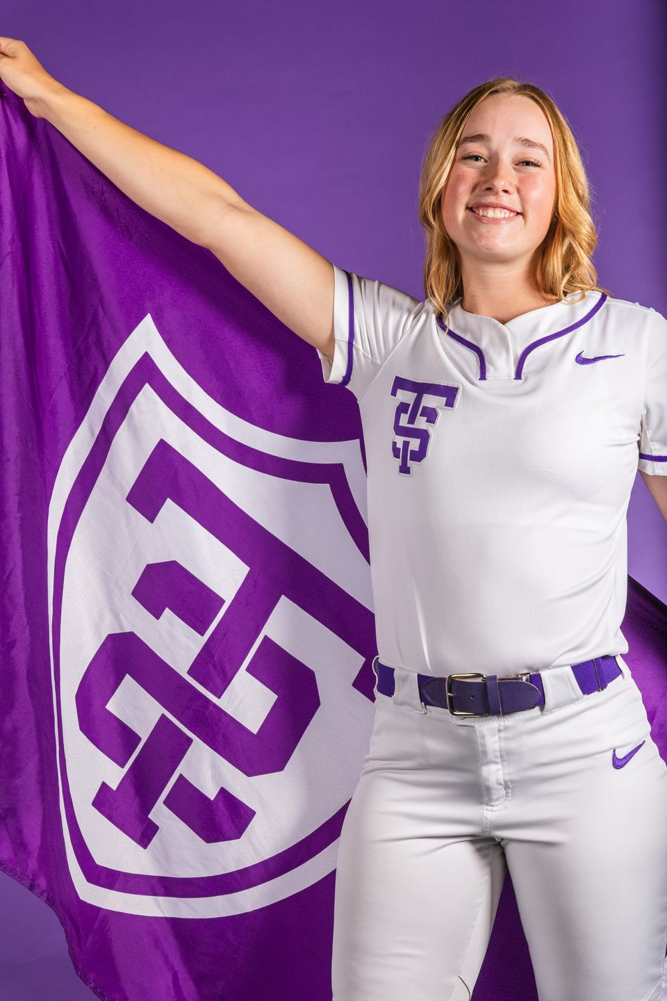 A studio portrait of Izzy Vick in a softball uniform, holding a St. Thomas flag behind her back 