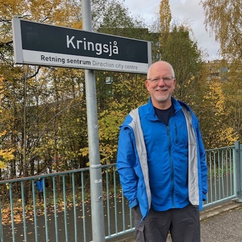 A man with a beard stands in front of a sign with words in a Scandinavian language